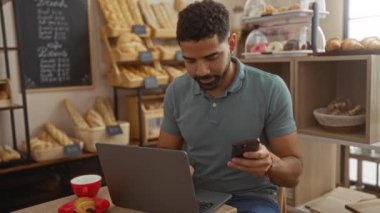 Genç adam ekmek ve pastalarla fırında dizüstü bilgisayar ve telefonla çalışıyor.