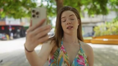 Genç bir kadın şehir manzarası ve yeşilliklerle çevrili canlı bir şehir parkında akıllı bir telefonla video görüşmesi yapıyor.