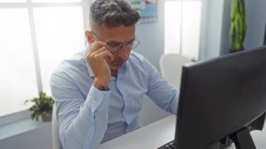 Ofiste gözlerini ovuşturan bir adam bilgisayar başında çalışırken yorgunluk ve stres belirtileri gösteriyor..