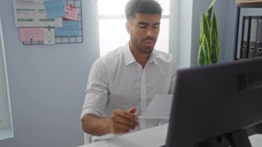 Genç adam ofis masasında belge okuyor, gözlükleri ayarlıyor, odaklanmış ifade, iş yeri, kapalı alan, gömlek, bilgisayar, iş yeri, evrak işleri.