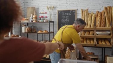 Pastanede çalışan bir adam, çeşitli hamur işleri ve somunlarla dolu kapalı bir dükkânda ekmek satan bir kadına yardım ediyor.