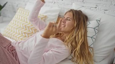 Genç bir kadın rahat bir yatak odasında uzanıyor, rahat pembe pijamalar giyiyor ve elleriyle anlamlı jestler yapıyor..