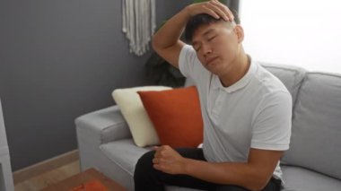 Beyaz gömlekli genç adam, modern bir oturma odasında gri bir koltukta oturuyor, ağrılı ya da rahatsız gibi görünürken başına masaj yapıyor, rahat ev dekoru ve minimalist mobilyalarla çevrili..