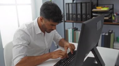 Ofiste çalışan, klavyeyle yazı yazan, boynunu esneten, yoğun bir iş yerinde stres ve yorgunluk belirtileri gösteren genç bir adam.