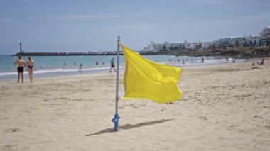 Lanzarote 'deki kumlu sahilde sarı bayrak, gündüz vakti açık mavi gökyüzünün altında yüzen ve kıyıda yürüyen insanlarla dolu kanarya adaları..