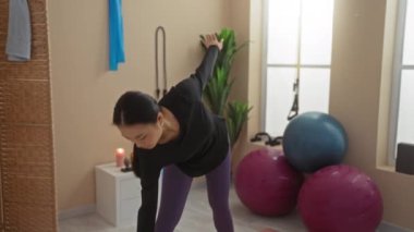 Spor salonunda spor toplarıyla esneyen, siyah bluz ve mor tayt giyen, esnekliğe odaklanan, spor aletleriyle çevrili bir kadın.