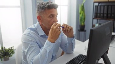 Orta yaşlı bir adam ofis odasında bilgisayar başında çalışırken modern iç mimari ve fabrikalarla kafa yoruyor.