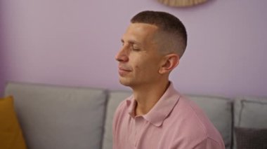 Oturma odasında rahat eden, pembe gömlek giyen, arka planda duvar dekoru ve minderleri olan mor renkli bir duvara yaslanmış genç bir adam.