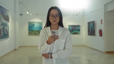 Beyaz gömlekli kadın sanat galerisinde akıllı telefon kullanıyor, teknoloji ve kültüre odaklanıyor.