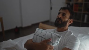 Yatakta sakallı genç bir adam ve loş ışıklı yatak odasında yastığa sarılıyor.