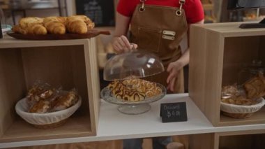 Pastanede çalışan, yeni pişmiş kruvasanları düzenleyen, kahverengi önlük ve kırmızı gömlek giyen, arka planda ekmek ve hamur işleri olan bir kadın..