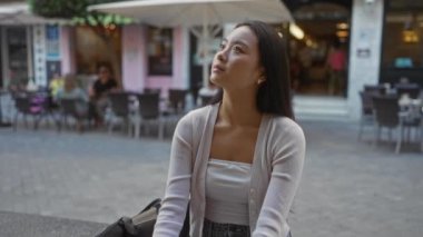 Şehir merkezinde bir kafede oturan genç bir kadın, düşünceli ve çevresindeki sokak hayatıyla meşgul görünüyor, sakin ve içsel bir anı öneriyor..