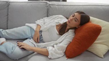 Rahat bir evde, rahat bir oturma odasında, rahat bir kanepede dinlenen, günlük kıyafetler giyen ve akıllı bir telefon tutan, gözleri kapalı rahat görünen bir kadın..