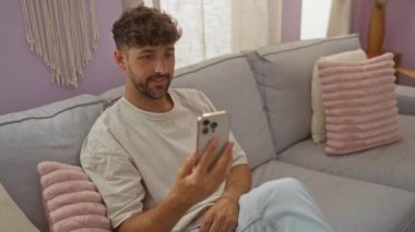İçerideki genç adam şık dekorlu rahat bir oturma odasında rahat bir kanepede otururken akıllı telefondan video sohbeti yapıyor..