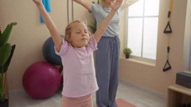 Anne ve kızı spor salonunda birlikte yoga yapıyorlar. Spor topları ve spor aletleri görünür.