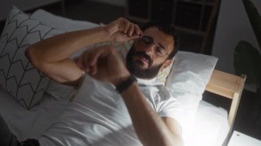 Beyaz tişörtlü, sakallı genç bir İspanyol gece yatağında gözlük çıkartıyor..