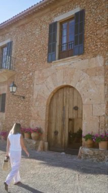 Güneşli bir günde, açık havada, Mallorca, İspanya 'da, büyük ahşap kapılı ve saksısı olan kırsal taş binaya doğru yürüyen bir kadın.