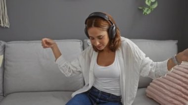 Kanepede dans eden, kulaklık takan, rahat bir oturma odasında müzik dinleyen bir kadın.