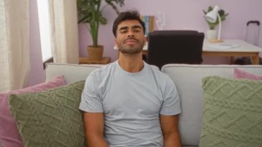 Oturma odasında oturan yakışıklı genç adam yastıklı bir kanepeden kameraya gülümseyip işaret ediyor ve arka planda bir masa var.