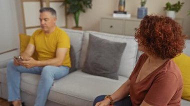 Evde orta yaşlı bir kadın ve erkek, kadın düşünceli görünürken, erkek telefonunu kullanıyor, rahat bir oturma odasında oturuyor, modern dekor ve tarafsız ses tonlarıyla.