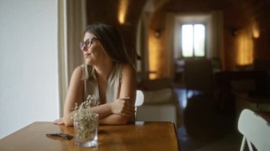 Kapalı mekanda gülümseyen gözlüklü genç bir kadın. Yumuşak pencere ışığı ve masanın üstünde vazosu olan rahat bir restoranda.