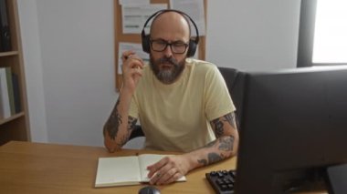 Sakallı ve dövmeli İspanyol bir adam, kulaklık takıyor, ofiste çalışıyor, notlar alıyor, ciddi ifadeler alıyor, bilgisayar, not defteri, odaklanıyor, merak ediyor, işyerinde çalışıyor.