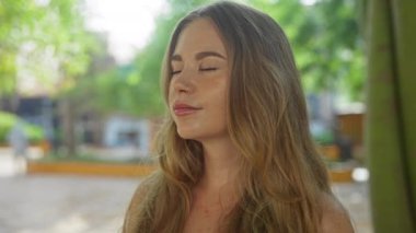 Sarı saçlı genç bir kadın güneşli bir günde yeşil arka planda bir şehir parkında gülümsüyor.