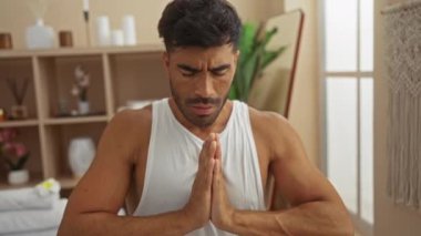 Huzurlu bir spa odasında meditasyon yapan bir adam. Elleri katlı ve gözleri kapalı. Etrafı sağlık dekoruyla çevrili.