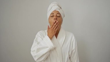Havlu ve bornoz giymiş orta yaşlı bir kadın yorulmadan esniyor, uykusuzluk ve bıkkınlık gösteriyor - beyaz arka planda izole edilmiş, yorgunluk nedeniyle ağızla ağzını kapatıyor.