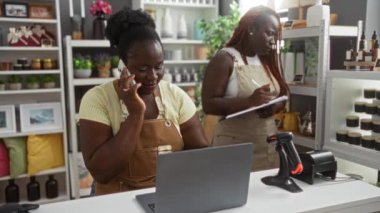 Biri telefonla konuşan, diğeri de dekoratif eşyalar ve bitkilerle çevrili notlar alan ev dekorasyonu mağazasında çalışan siyahi kadınlar..