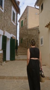 Valldemossa, Mallorca, İspanya 'nın dar taşlı caddelerinde yürüyen bir kadın hasır bir çanta taşıyor ve güneşli bir günde siyah bir elbise giyiyor.