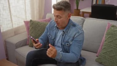 Orta yaşlı bir adam rahat bir oturma odasında kot ceket ve kot pantolon giyerken renkli yastıklarla bir koltukta kutlama yapıyor..