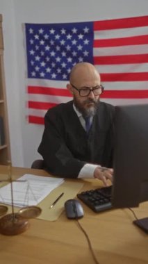 Arka planda Amerikan bayrağı olan, takım elbiseli ve gözlüklü, bilgisayarda yasal evraklar ve terazilerle çalışan bir adam..