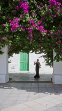 Haria, Lanzarote, Kanarya Adaları 'ndaki yeşil bir kapının yanında pembe çiçeklerin altında yürüyen bir kadın canlı renkleri ve yaz havasını gözler önüne seriyor.