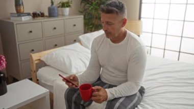 Orta yaşlı, pijamalı, yatakta oturan, telefon kullanan ve rahat yatak odasında kahve içen bir adam..