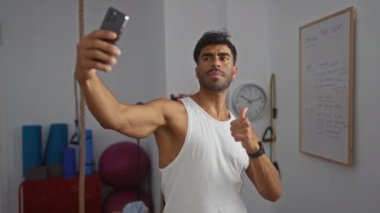 Genç adam spor salonunda özçekim yapıyor. Arka planda beyaz tahta ve egzersiz ekipmanları var.