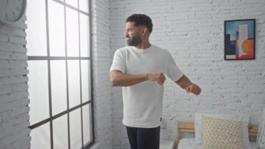 Yatak odasında duran genç adam beyaz gömlek ve donanma pantolonu içinde doğal ışıkla aydınlatılmış beyaz tuğla duvarlar ve minimalist dekorlarla çevrili büyük pencerenin yanında duruyor.