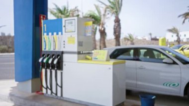 Benzin pompalı bir benzin istasyonunun bulanık, bulanık manzarası, yanına park edilmiş beyaz bir araba ve güneşli bir günde arka planda palmiye ağaçları..