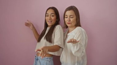 Anne ve kızı pembe bir duvara karşı gülümseyip dans ediyorlar. Sevgi, neşe ve aile bağlarını ifade ediyorlar.