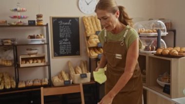 Kahve fincanı tutarken pastanedeki kumaşlı masayı temizleyen kadın, dükkanın içinde taze ekmek somunları ve hamur işleriyle çevrili..