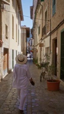 Fornalutx, Mallorca 'da dar bir kaldırım taşı caddesinde yürüyen, elinde örülmüş bir çanta, beyaz elbise ve şapka giyen, güneşli bir günde kırsal Akdeniz mimarisi ile çevrili bir kadın.
