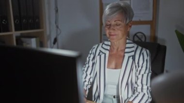 Ofiste çalışan gri saçlı bir kadın, bilgisayar başında çalışıyor, çizgili ceket giyiyor, ekrana odaklanıyor, uçakta belgeler, arka planda kitap rafları, iç dekorasyon..