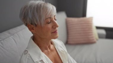 Kısa gri saçlı, rahat bir oturma odasındaki kanepede dinlenen sakin bir kadın.