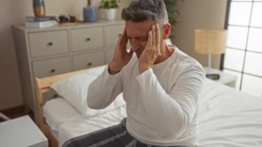 Orta yaşlı bir adam yatak odasında baş ağrısı olan bir yatakta oturuyor. Şakaklarını rahatsızlık içinde tutuyor. Etrafı mobilya ve bitkilerle çevrili.