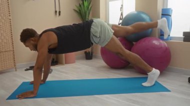 Spor merkezindeki bir paspasın üzerinde yoga yapan genç bir adam, spor aletleri ve arka planda egzersiz topları var..