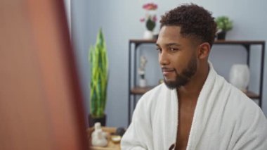 Sağlık merkezindeki genç adam beyaz bir bornoz giyerken gülümsüyor ve elinde arka planda bitki ve raflarla bambu bir diş fırçası tutuyor..