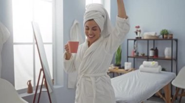 Bir sağlık merkezinde elinde bardakla mutlu bir şekilde dans eden bir kadın. Üzerinde bornoz ve havluyla güzel ve rahat bir iç mekanda dans ediyor..