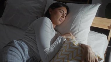 Yatak odasında beyaz gömlek ve pijama giymiş bir yastıkla uzanan bir kadın evin içinde rahatsız görünüyor.