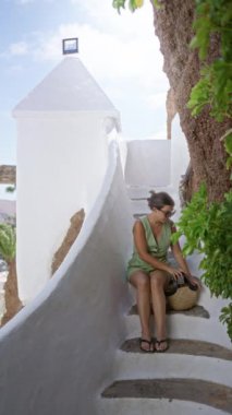 Yeşil elbiseli kadın lagomar, lanzarotta beyaz basamaklarda oturuyor, kırsal beyaz mimari ve yeşillikle süslenmiş, güneşli bir gökyüzünün altında huzurlu bir tatil ortamı yayıyor.