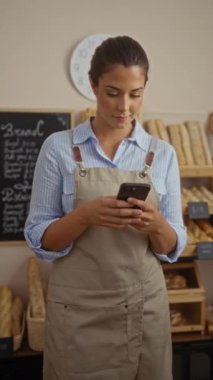 Fırında akıllı telefon kullanan genç bir kadın, arka planda ekmek somunları olan bir önlük giyiyor.